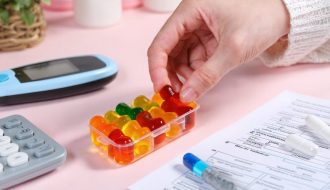 A hand holds a vibrant HHC gummy above a pill organizer, symbolizing conscious integration of these treats into a diabetes management routine, with tools and notes scattered around.