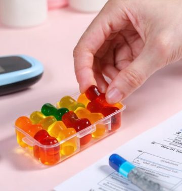 A hand holds a vibrant HHC gummy above a pill organizer, symbolizing conscious integration of these treats into a diabetes management routine, with tools and notes scattered around.