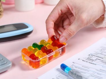 A hand holds a vibrant HHC gummy above a pill organizer, symbolizing conscious integration of these treats into a diabetes management routine, with tools and notes scattered around.